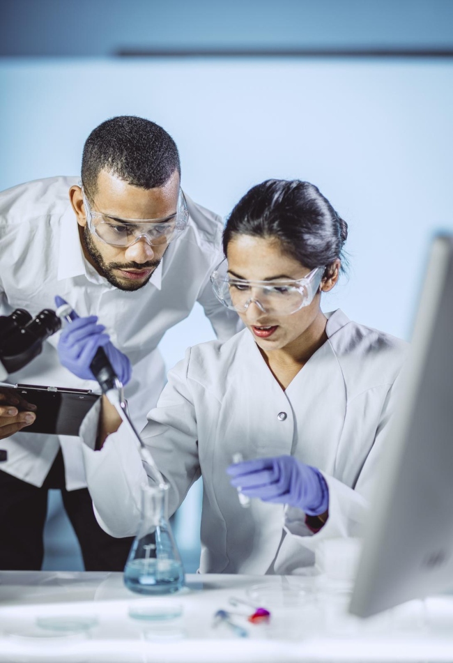 Two scientists wearing personal protective equipment (i.e. goggles, gloves, and laboratory coats) are stationed behind a laboratory work bench with a computer on top. One scientist is pipetting liquid into a flask, while the other observes.